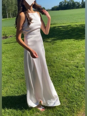 Ljusrosa balklänning Bubbleroom - Elegant vit långklänning i satin med ärmlös design och rund halsringning. Klänningen har en mjukt draperad rygg och slät, figurnära passform som faller snyggt längs kroppen. Perfekt för speciella tillfällen och student.