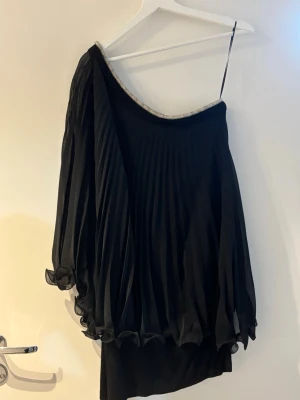 Svart one shoulder klänning med volang - Snygg svart kortklänning med plisserad, luftig ärm och one shoulder-design. Klänningen har en glittrig kant vid halsen och volangdetaljer längst ner på ärmen. Perfekt för dig som vill sticka ut med en unik och trendig look. S-M storlek 