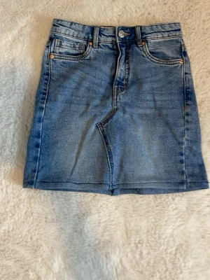 Blå jeanskjol med klassisk passform - Snygg blå jeanskjolmed klassisk femficksdesign och knapp- samt dragkedjestängning framtill. Kjolen har en rak, kort modell och är tillverkad i denim med tydliga sömmar och bälteshällor. Perfekt för en avslappnad och trendig look. Sytt själv. Storlek 146.