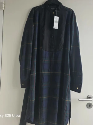 Mörkblå rutig långklänning från ralph lauren  - Snygg långklänning från Rodebjer i mörkblått rutigt tyg med svarta detaljer framtill. Klänningen har lång ärm, knappar vid ärmslut och en unik svart panel över bröstet. Perfekt för dig som gillar en avslappnad men ändå stilren look.