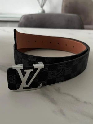  bälte med spänne - Snyggt bälte från med klassiskt rutigt mönster i svart och grått. Bältet har ett stort spänne i silverfärg och är gjort i skinn. Perfekt accessoar för att lyfta din outfit.