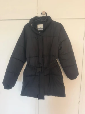 Svart pufferjacka med bälte från NA-KD - Snygg svart pufferjacka från NA-KD med hög krage och avtagbart midjebälte. Jackan är vadderad och har långa ärmar samt dold dragkedja och tryckknappar framtill. Perfekt för kalla vinterdagar och ger en trendig siluett.