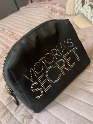 Svart necessär Victoria's Secret - Snygg svart necessär från Victoria's Secret med glittrande stenar som bildar loggan på framsidan. Väskan har dragkedja med gulddetaljer och är tillverkad i ett mjukt tygmaterial. Perfekt för smink eller småprylar.