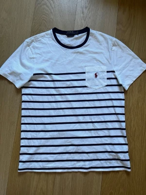 Randig t-shirt från Polo Ralph Lauren - Tjena! Säljer nu min vita t-shirt Polo Ralph Lauren som aldrig lämnat garderoben pga storleken. T-shirten har mörkblå horisontella ränder, en bröstficka med röd broderad logga och en rund halsringning med mörkblå kant. Materialet är mjuk bomull, väldigt skönt. Hör av dig vid minsta fundering!