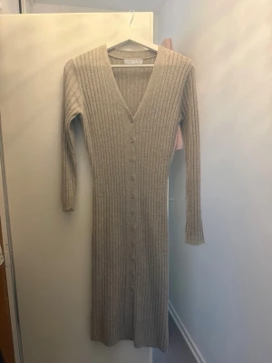 Beige ribbad stickad klänning - Långärmad beige klänning i ribbstickat material med knappar längs hela framsidan och djup V-ringning. Klänningen är kroppsnära och har en mjuk, stretchig känsla. Perfekt för dig som gillar en enkel men snygg look.