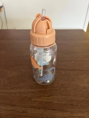 Vattenflaska med dinosauriemotiv - Transparent vattenflaska med sugrör och persikofärgat lock. Flaskan är dekorerad med söta dinosaurier i blått och orange. Perfekt för att ha med sig till gymmet, skolan eller utflykten. Tillverkad i plast.