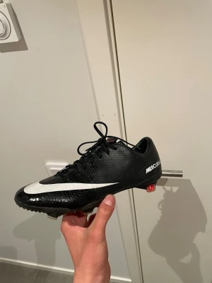 Nike Mercurial fotbollssko svart - Säljer en svart Nike Mercurial fotbollssko med vit swoosh och text på sidan. Skon har en låg profil, snörning och mönstrad yta i syntetmaterial. Dobbar i plast och metall för bra grepp på planen. Perfekt för dig som vill ha snabbhet och kontroll.