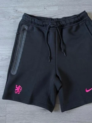 Svarta Nike shorts med rosa detaljer - Svarta shorts från Nike med rosa logga och lejonmärke. De har snörning i midjan och en dragkedjeficka på sidan. Perfekta för en sportig look och tillverkade i mjukt material.