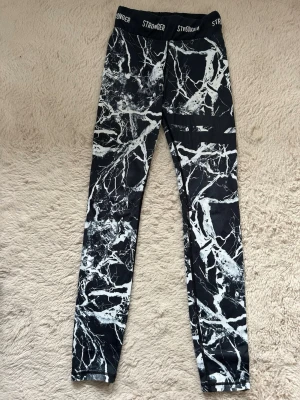Svarta leggings med marmormönster Stronger - Snygga svarta leggings från Stronger med vitt marmormönster och hög midja. Perfekta för träning. Stretchigt material som sitter skönt och följer kroppen.