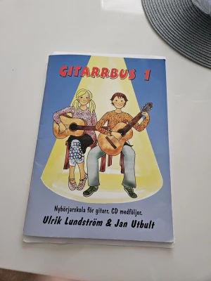 Gitarrbus 1 av Lundström & Utbult - Gitarrbus 1 är en nybörjarskola för gitarr med färgglad framsida där två personer spelar gitarr. Boken har blått omslag med röd och gul text. . Perfekt för dig som vill lära dig spela gitarr från grunden. Har tappat CdD