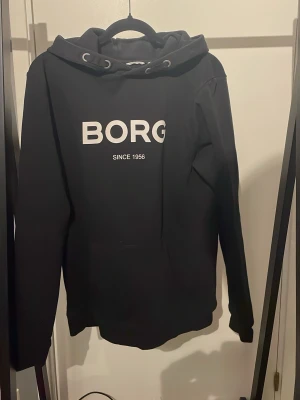 Svart hoodie från Björn Borg - Snygg svart hoodie från Björn Borg med stor vit logga 'BORG' och texten 'SINCE 1956' på bröstet. Klassisk modell med huva och dragsko, samt ribbade muddar vid ärmslut och nederkant. Perfekt för en chill och sportig look.