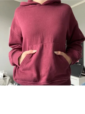Vinröd hoodie med magficka - Enkel och snygg vinröd hoodie med huva och stor magficka framtill. Hoodien har ribbade muddar vid ärmslut och nederkant. Perfekt för en avslappnad och trendig stil. Materialet känns mjukt och bekvämt mot huden.