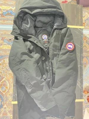 Svart dunjacka från Canada Goose - Säljer en svart dunjacka från Canada Goose med huva och klassisk logotyp på ärmen. Jackan har dragkedja och knappar framtill samt en varm, vadderad insida. Perfekt för kalla vinterdagar och riktigt snygg streetstil.