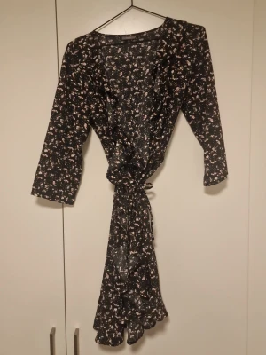 Svart blommig omlottklänning - Svart kort omlottklänning med små blommor i rosa och vitt. Klänningen har trekvartsärm, knyt i midjan och volangkant nertill. Perfekt för dig som gillar en feminin och avslappnad stil.