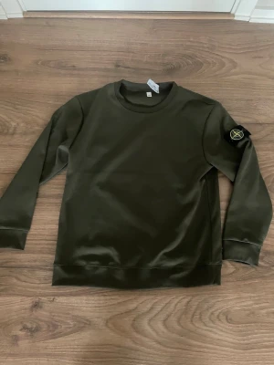Olivgrön sweatshirt från Stone Island - Snygg olivgrön sweatshirt från Stone Island med klassisk rund hals och ribbade muddar. Tröjan har den ikoniska Stone Island-loggan på vänster ärm och är tillverkad i mjukt bomullsmaterial. Perfekt för en avslappnad och trendig stil.