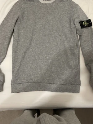 Grå sweatshirt från Stone Island - Snygg grå sweatshirt från Stone Island med klassisk rund hals och ribbade muddar. Tröjan har den ikoniska loggan på vänster ärm och är tillverkad i mjukt bomullsmaterial. Perfekt för en avslappnad och stilren look.