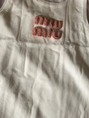 Vit ribbad linnetopp från Miu Miu - Superfin vit ribbad linnetopp från Miu Miu med rosa pärlbroderade bokstäver framtill. Toppen är ärmlös och har en klassisk rund halsringning. Perfekt att styla med jeans eller kjol för en trendig look.