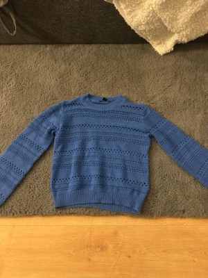 Blå stickad tröja med hålmönster - Säljer en blå stickad tröja med snyggt hålmönster och rund hals. Tröjan har långa ärmar och ribbade muddar vid ärmslut och nederkant. Perfekt för dig som vill ha en trendig och bekväm look.
