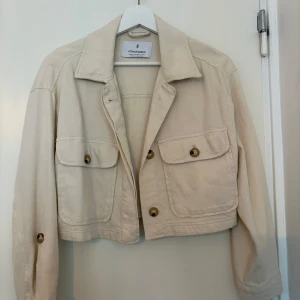 Beige croppad trenchcoat från Stradivarius - Stilren beige croppad overshirt från Stradivarius med två stora bröstfickor och knappar framtill. Jackan har lång ärm med knappdetalj och klassisk krage. 