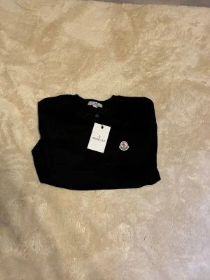 Svart sweatshirt från Moncler - Svart sweatshirt från Moncler med rund halsringning och Moncler-logga broderad på bröstet. Tröjan har en klassisk passform och är tillverkad i mjukt material som känns skönt mot huden. Perfekt för en stilren och avslappnad look.