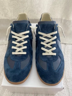 Blå Maison Margiela sneakers - Snygga sneakers från Maison Margiela i blå mocka och läder med vita skosnören och klassisk gummisula. Skorna har en låg silhuett och stilren design som passar perfekt till streetwear eller casual outfits.