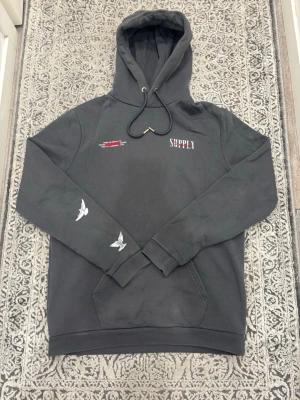 Grå hoodie från Supply & Demand - Grå hoodie med tryck från Supply & Demand. Framsidan har små logotyper och text, medan baksidan har en stor ängel med röda detaljer och vita duvor. Fickor framtill och dragsko i huvan. Snygg streetwear-stil med unika detaljer.