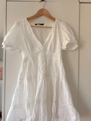 Vit sommarklänning med puffärm - Söt vit klänning med v-ringning och puffärmar. Klänningen har broderade detaljer och smock i midjan för en skön passform. Tillverkad i ett lätt och luftigt tyg, perfekt för varma dagar. Kjolen är volangig och ger en romantisk känsla.