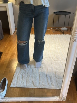 Blå raka jeans med slitningar - Säljer ett par blå raka jeans med stora slitningar över knäna. Jeansen har hög midja.