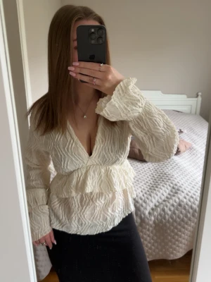 Vit volangblus från Gina Tricot - Säljer en superfin vit blus från Gina Tricot med volangdetaljer och djup V-ringning. Blusen har ett krinklat tyg som ger en cool struktur och långa ärmar med lätt utsvängda avslut. Perfekt att styla med både jeans och kjol!