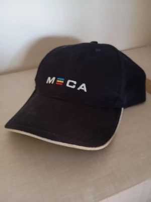 Svart keps från MECA med brodyr - Svart keps med böjd skärm och vit kant. Framtill finns broderad MECA-logga i vitt med färgglada ränder. Klassisk sexpanelsdesign och justerbar baksida. Perfekt accessoar för en sportig och avslappnad stil.
