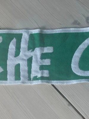 Grön fotbollshalsduk Elche CF - Snygg grön halsduk med Elche CF:s klubbmärke och vit text. Perfekt accessoar för dig som vill visa ditt stöd för laget. Halsduken har vita fransar i ändarna och är tillverkad i ett mjukt material som passar bra till matchdagar.