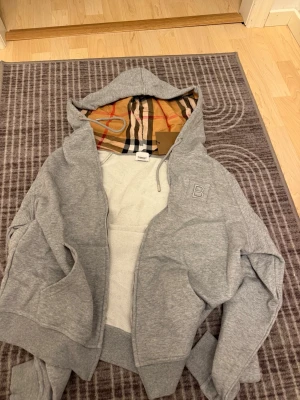 Grå hoodie från Burberry med huva - Snygg grå hoodie från Burberry med dragkedja och klassiskt rutigt foder i huvan. Hoodien har broderat B på bröstet, ribbade muddar och fickor framtill. Perfekt för en avslappnad och trendig stil.