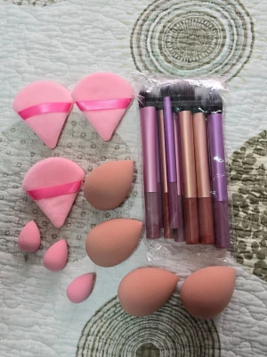 Set med sminkborstar och svampar - Ett komplett kit med sminkborstar i metallicrosa och lila toner samt flera makeupsvampar i olika storlekar och former. Borstarna har mjuka syntetiska borst och handtag i metall. Svamparna är i rosa och beige nyanser, perfekta för foundation, concealer och puder.