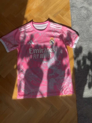 Rosa Real Madrid matchtröja Adidas - Säljer en rosa Real Madrid matchtröja från Adidas med vita detaljer och drakmönster. Tröjan har klubbmärke, Adidas-logga och Emirates Fly Better-tryck på bröstet. Perfekt för dig som vill sticka ut på planen eller läktaren.