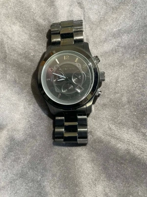 Svart klocka från Michael Kors - Snygg svart klocka från Michael Kors med metallarmband och rund urtavla. Klockan har flera små urtavlor och tydliga siffror. Perfekt accessoar för dig som gillar stilrena detaljer och en modern look.