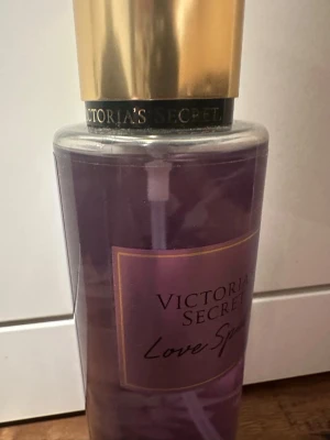Victoria's Secret Love Spell mist - Victoria's Secret Love Spell body mist i en genomskinlig plastflaska med lila vätska och guldfärgat lock. Doften har inslag av körsbärsblom och persika. Flaskan har en elegant och feminin design med svart och guld detaljer. Aldrig använts. 
