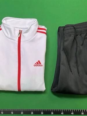 Adidas träningsset med röd/vit jacka - Snyggt träningsset från Adidas med en vit jacka med röda detaljer och dragkedja samt svarta byxor med vita ränder. Jackan har hög krage och Adidas-logga på bröstet. Byxorna har dragsko i midjan och klassiska Adidas-ränder längs benen. Perfekt för träning eller chill.