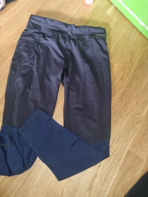 Svarta träningstights från Newbody - Säljer ett par svarta tights från Newbody med hög midja och praktisk ficka på sidan. Tillverkade i stretchigt och mjukt material som sitter skönt på kroppen. Perfekta för träning eller chill. Snygg och enkel design med diskret logga vid midjan.