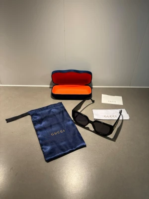 Svarta solglasögon  - Snygga svarta solglasögon med rektangulär form och breda skalmar. På sidan sitter den klassiska i guld. Kommer med blå sammetsfodral, putsduk och förvaringspåse. Perfekt accessoar för att lyfta din stil.