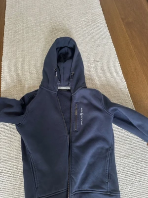 Mörkblå hoodie från Sail Racing - Mörkblå hoodie från Sail Racing med dragkedja framtill, justerbar huva och en bröstficka med dragkedja. Tröjan har ribbade muddar vid ärmsluten och två sidofickor. Perfekt för dig som gillar sportig och stilren look.