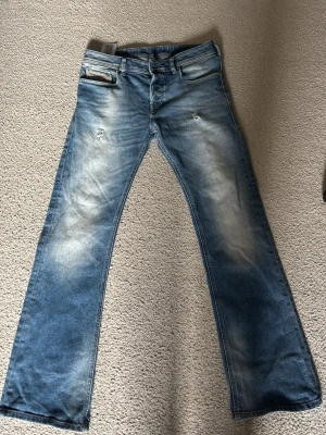 Blå bootcut jeans från Diesel - W28 men sitter som 29 och L är 30 dock långa Säljer ett par blå bootcut jeans från Diesel med slitningar och ljusa tvättade partier. Jeansen har klassisk femficksmodell, låg midja och detaljerade sömmar. Materialet är denim i bomull och passformen är avslappnad med utsvängda ben.