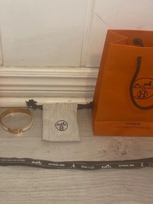 H&M vit och guld armband - Säljer ett exklusivt armband från H&M i vit emalj med guldfärgade detaljer och klassiskt H-spänne. Armbandet kommer med originalpåse och H&M presentpåse, perfekt för dig som gillar lyxiga accessoarer och ikoniska designer.