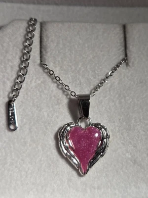 Halsband med lila hjärthänge - Snyggt handgjort halsband med ett glittrigt lila hjärthänge omslutet av silverfärgade vingar som lyser lila i mörker efter att ha varit i dagsljus eller solljus. Kedjan är i silverfärgad rostfritt stål och har justerbar längd. Perfekt för dig som gillar unika och färgglada accessoarer med lite extra detalj. 