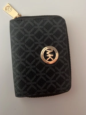 Svart plånbok från Michael Kors - Stilren svart plånbok från Michael Kors med guldfärgad logotyp på framsidan och mönstrad yta. Plånboken har dragkedja runtom och flera fack för kort. Materialet är konstläder och detaljerna är i metall. (EJ ÄKTA)