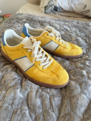 Gula sneakers med blå och vita detaljer - Snygga sneakers i gult tyg och mocka med vita och blå detaljer. Skorna har vita snören, vit sula och en brun yttersula. Perfekt för dig som vill sticka ut med färg! Klassisk låg modell som passar till jeans eller shorts.