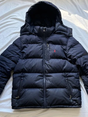 Marinblå dunjacka med huva från Polo Ralph Lauren - Det är en gorham Snygg marinblå jacka från Polo Ralph Lauren med stor huva och dragkedja framtill som kostar ordinarie ny pris 5400 kr. Jackan har två sidofickor med dragkedja och en bröstficka med röd logga. Perfekt för kalla dagar och riktigt skön att bära. Har några små slitningar men syns inte mycket när man har på den.