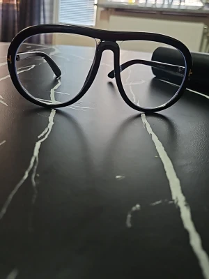 Svarta Tom Ford pilotbågar - Snygga svarta solglasögon från Tom Ford med klassisk pilotform och tydliga bågar. Glasögonen har en stilren design med diskreta gulddetaljer vid gångjärnen. Perfekta för dig som vill ha en trendig accessoar med exklusiv känsla.