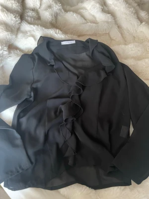 Svart volangblus från Jasmin - Stilren svart blus från Jasmin med långa ärmar och snygga volangdetaljer längs halsringningen och framtill. Tillverkad i ett tunt, lätt material, något genomskinlig. Storlek M, aldrig använd🩷