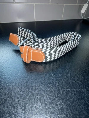 Flätat bälte - Snyggt svartvitt bälte med zigzag-randigt mönster och bruna läderdetaljer vid spännet. Perfekt accessoar för att lyfta din outfit och ge en cool touch. Justerbar längd och passar till många olika stilar.