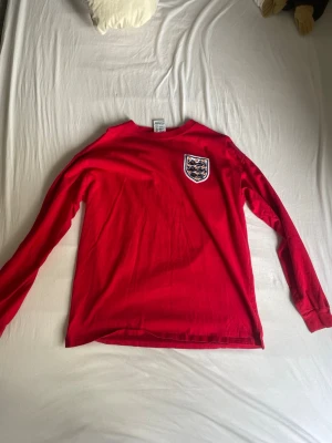 Långärmad England tröja  - Säljer en röd långärmad tröja med England-emblem på bröstet. Tröjan har rund hals och är tillverkad i mjukt material, perfekt för dig som gillar sportiga och klassiska plagg. Emblemet har detaljer i vitt och svart.